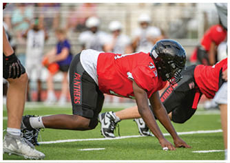 An adult in a red football jersey and black helmet is crouched on a football field in a ready position with other players in similar uniforms in the background. Description generated by AI
