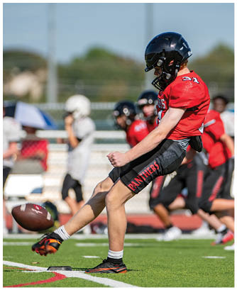 A person wearing a red jersey and black helmet is kicking a football on a green field with other players and spectators blurred in the background. Description generated by AI