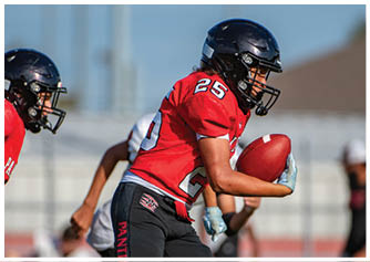 A person wearing a red football jersey and black helmet is holding a football and appears to be running on a field with another player in the background. Description generated by AI