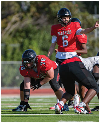 People in red and white football uniforms are positioned on a field preparing for a play, with one person in red signaling with their hand. Description generated by AI