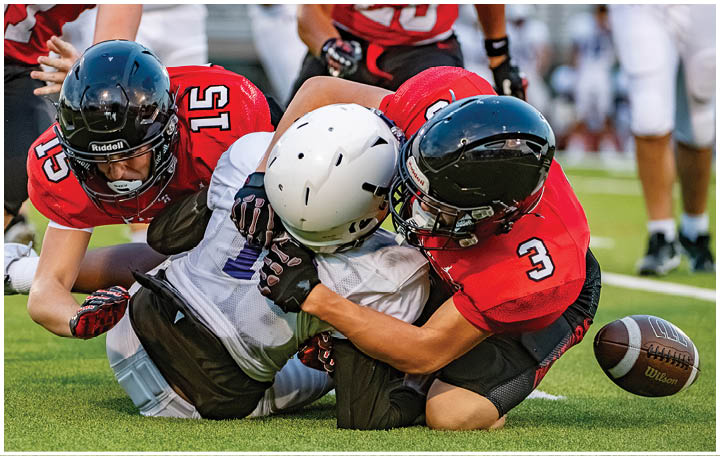 Two people in football gear are on the ground with one tackling the other while a third person reaches toward them and a football is rolling nearby on the grass field. Description generated by AI