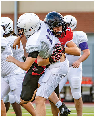 Two people wearing football helmets and uniforms are engaged in a tackle during a game, with one person in a red jersey and the other in a white jersey. Description generated by AI