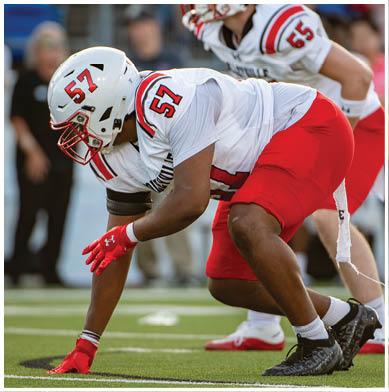 Two people in football uniforms with white helmets and red pants are crouched in a ready position on a green field during a game. Description generated by AI