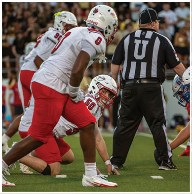 Several people in football uniforms and helmets are positioned on a field with a referee standing nearby. Description generated by AI