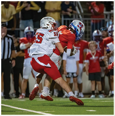 Two people in football uniforms are engaged in a tackle during a game, with one player in white and red attempting to stop another player in orange holding the ball. Description generated by AI