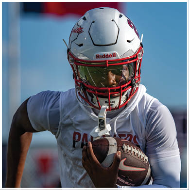 A person wearing a white football helmet with a red faceguard and a white sports jersey is holding a football while preparing to run. Description generated by AI