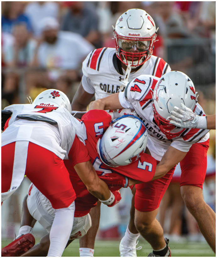 Four football players in red and white uniforms are engaged in a play on a green field with a blurred crowd in the background. Description generated by AI