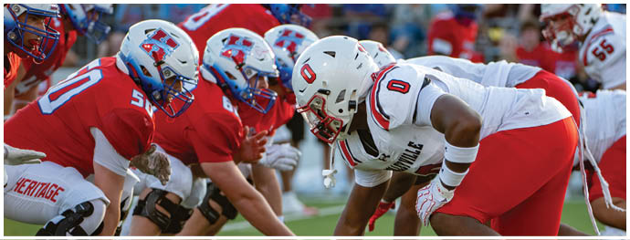 Two opposing American football teams in red and white uniforms are lined up facing each other on the field, preparing for a play. Description generated by AI
