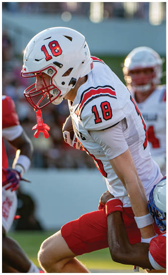 A person wearing a white football uniform with the number 18 on the helmet and jersey is leaning forward in a ready stance on a field. Description generated by AI