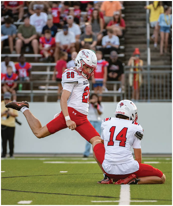 Two people in football uniforms with helmets are on a field, one holding the ball in place while the other prepares to kick it. Description generated by AI
