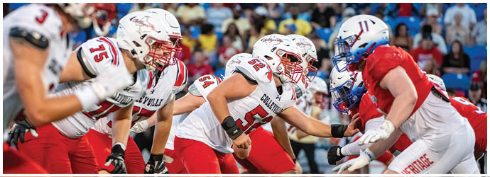 Two opposing American football teams in helmets and uniforms are lined up facing each other on the field with a crowd in the background. Description generated by AI