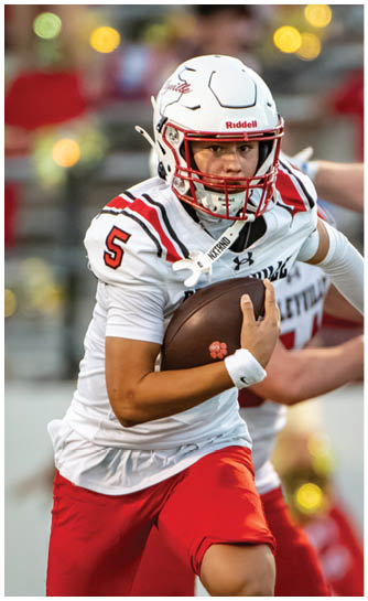 An adult wearing a white football helmet and jersey with red pants is running on a football field while holding a football, with other players and a blurred crowd in the background. Description generated by AI