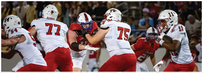 People wearing football uniforms are engaged in a play on a field with one person holding the ball and others blocking or preparing to receive. Description generated by AI