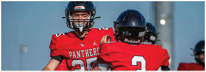 Two people wearing football helmets and red jerseys with “Panthers" on the front are engaged in a conversation or interaction on a field under a clear sky. Description generated by AI