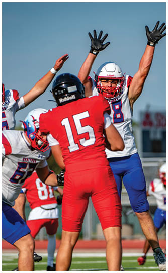 Four people in football uniforms are actively reaching and preparing to catch or block a football on a field during a game. Description generated by AI