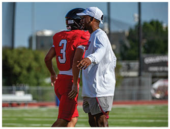 A person wearing a white long-sleeve shirt and cap is standing next to a person in a red football uniform with the number three on the back, on a sports field. Description generated by AI
