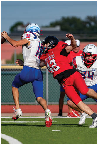 A person in a white and blue football uniform is throwing a football while being tackled by a person in a red football uniform on a green field with a coach in red watching in the background. Description generated by AI