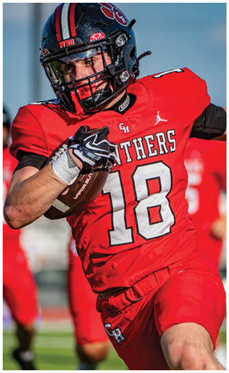A person wearing a red football uniform with the number 18 is running on a field while holding a football and evading a player in a white and blue uniform. Description generated by AI