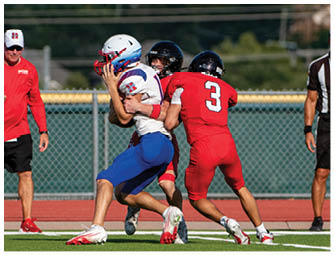 Two people in football uniforms are tackling a person in a white helmet and blue pants who is holding a football on a field with a coach and referee nearby. Description generated by AI