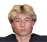 A person with medium-length blond hair wearing a dark sports jersey is looking directly at the camera with a neutral expression. Description generated by AI
