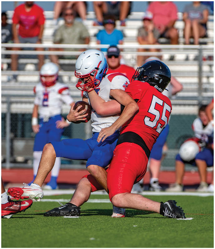 A person in a white helmet and blue pants is holding a football while being tackled by a person in a red uniform with the number 55 on it during a game on a green field with spectators in the background. Description generated by AI