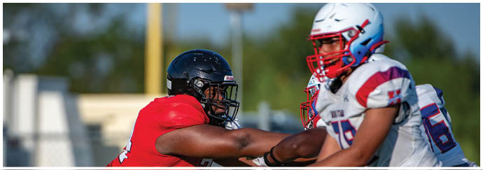 Two people in football uniforms are engaged in a physical play on a field, with one wearing a red jersey and black helmet and the other in a white jersey with red and blue accents. Description generated by AI