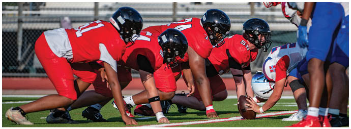 A group of people in red sports uniforms and helmets are lined up in a crouched position facing a person in a white and blue sports uniform on a football field with empty bleachers in the background. Description generated by AI