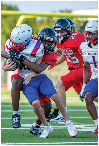 Two people in football uniforms and helmets are engaged in a tackle on a grassy field while two others in similar gear stand nearby. Description generated by AI