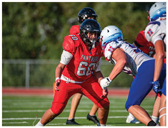 A person wearing a red football uniform with the word Panthers and number 68 is in a defensive stance facing an opponent in a white and blue football uniform on a grassy field. Description generated by AI