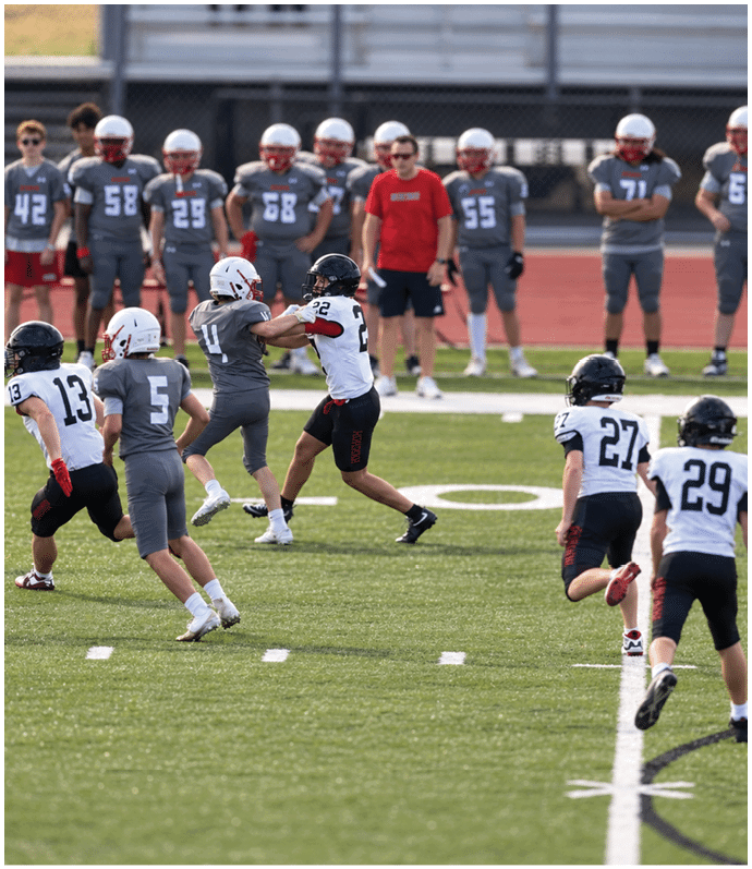 People in sports uniforms are playing football on a field with others standing on the sidelines in the background. Description generated by AI