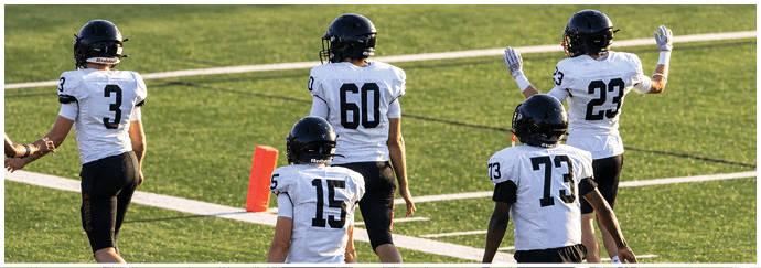 Several people wearing white sports jerseys and helmets are standing on a green field with white lines, appearing to prepare for or participate in a football game. Description generated by AI