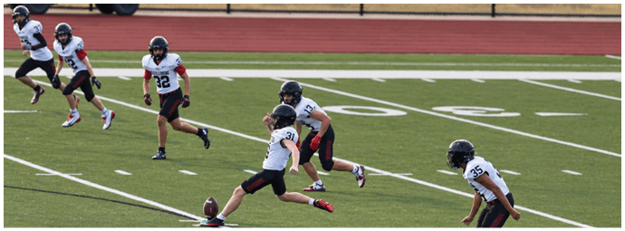 People in sports uniforms are playing football on a field with one person kicking the ball while others run nearby. Description generated by AI