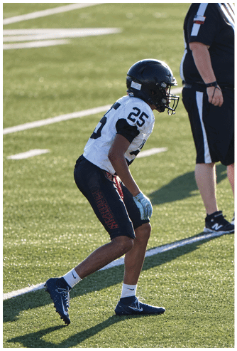 A person wearing a white football jersey with the number 25, black helmet, and gloves is standing on a grassy field. Description generated by AI