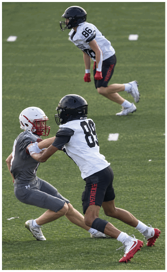 Two people wearing black helmets and white football jerseys with numbers 86 and 89 are on a grassy field, appearing to be engaged in a football game or practice. Description generated by AI