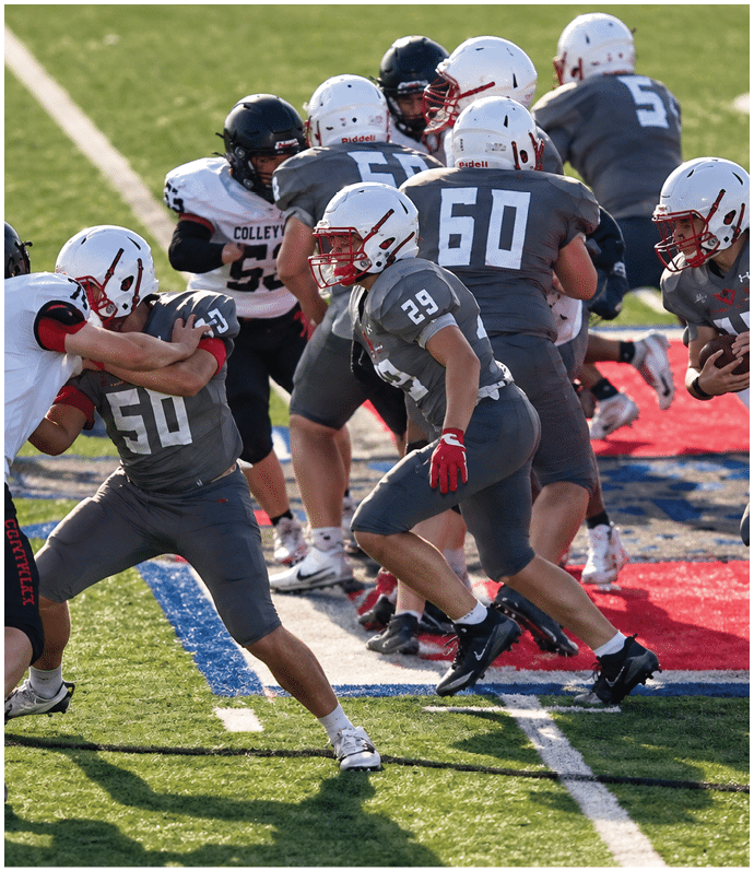 Players in gray and white football uniforms are engaged in a play on a green field with red and blue markings. Description generated by AI
