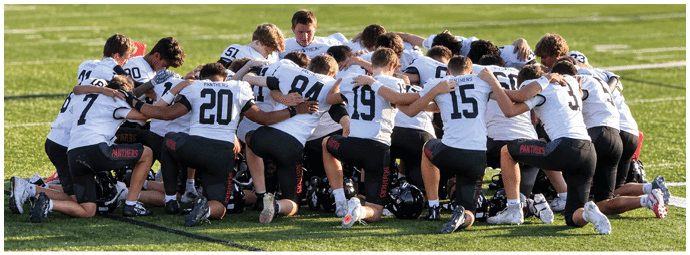 A group of people wearing sports uniforms are kneeling in a circle on a grassy field with their arms around each other. Description generated by AI