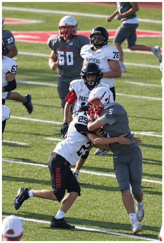 Players in football uniforms are actively engaged in a game on a grassy field with some players tackling and others running. Description generated by AI