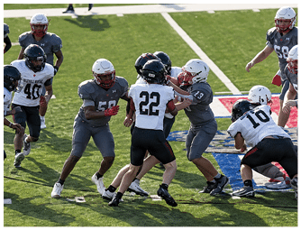 People wearing helmets and football uniforms are actively engaged in a play on a grassy field with yard lines and a colorful logo visible. Description generated by AI
