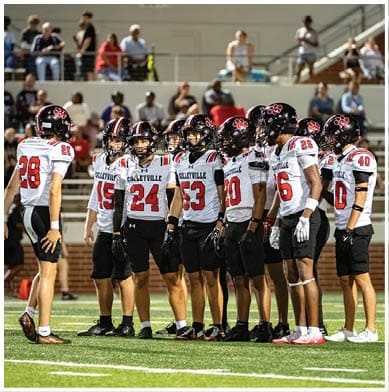 A group of people wearing football uniforms with helmets stand together on a field while others watch from the bleachers in the background. Description generated by AI