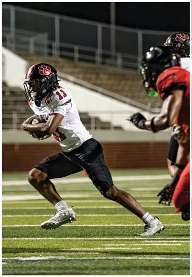 A person wearing a football uniform with the number 11 is running on a field while holding a football, with another player partially visible nearby. Description generated by AI