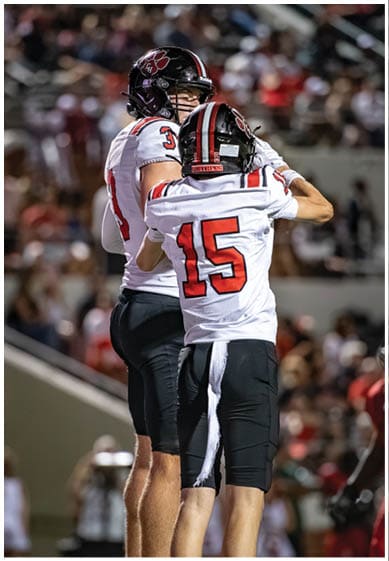 Two people in football uniforms are standing close together on a field, with one wearing a jersey numbered 15 and the other numbered 3. Description generated by AI