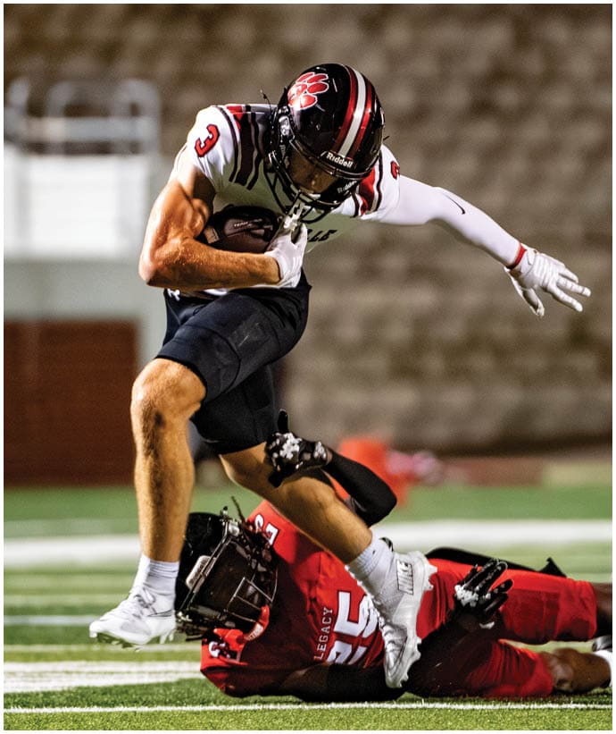A person wearing a football uniform with the number three is running with the ball while another person on the ground attempts to tackle them during a game. Description generated by AI
