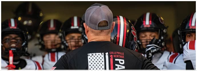 A person wearing a cap and a shirt with “Colleyville" on the back is facing a group of people in football helmets and uniforms. Description generated by AI
