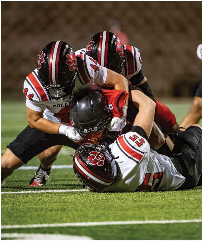 Three people in football uniforms are tackling a player in a red jersey on a grassy field during a game. Description generated by AI