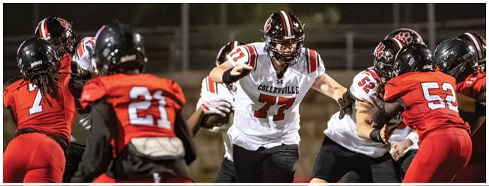 Adults wearing football uniforms are engaged in a game on a field with one player in a white jersey running toward the camera while others in red jerseys prepare to block or tackle. Description generated by AI