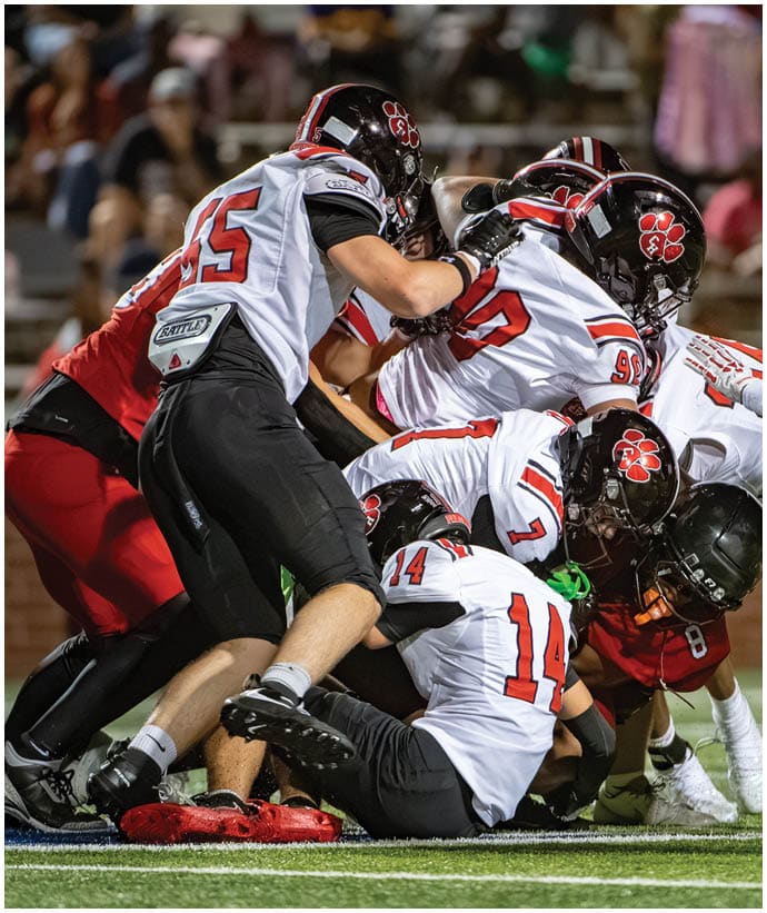 Several people wearing football helmets and uniforms are engaged in a close, intense play on a football field. Description generated by AI