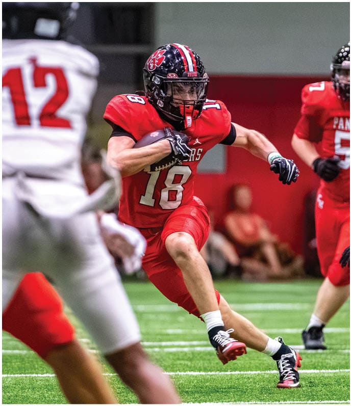 A person in a red football uniform is running with the ball while another person in a white uniform with the number 12 is in the foreground on a football field. Description generated by AI