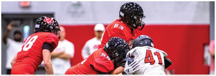 People wearing helmets and sports uniforms are engaged in a football game or practice indoors with a red and white background. Description generated by AI