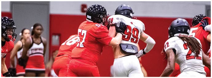 People in red and white football uniforms are engaged in a play on an indoor field with cheerleaders visible in the background. Description generated by AI