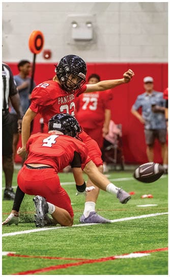 Two people in red football uniforms and helmets are on an indoor field with one person holding the ball on the ground and the other kicking the ball. Description generated by AI